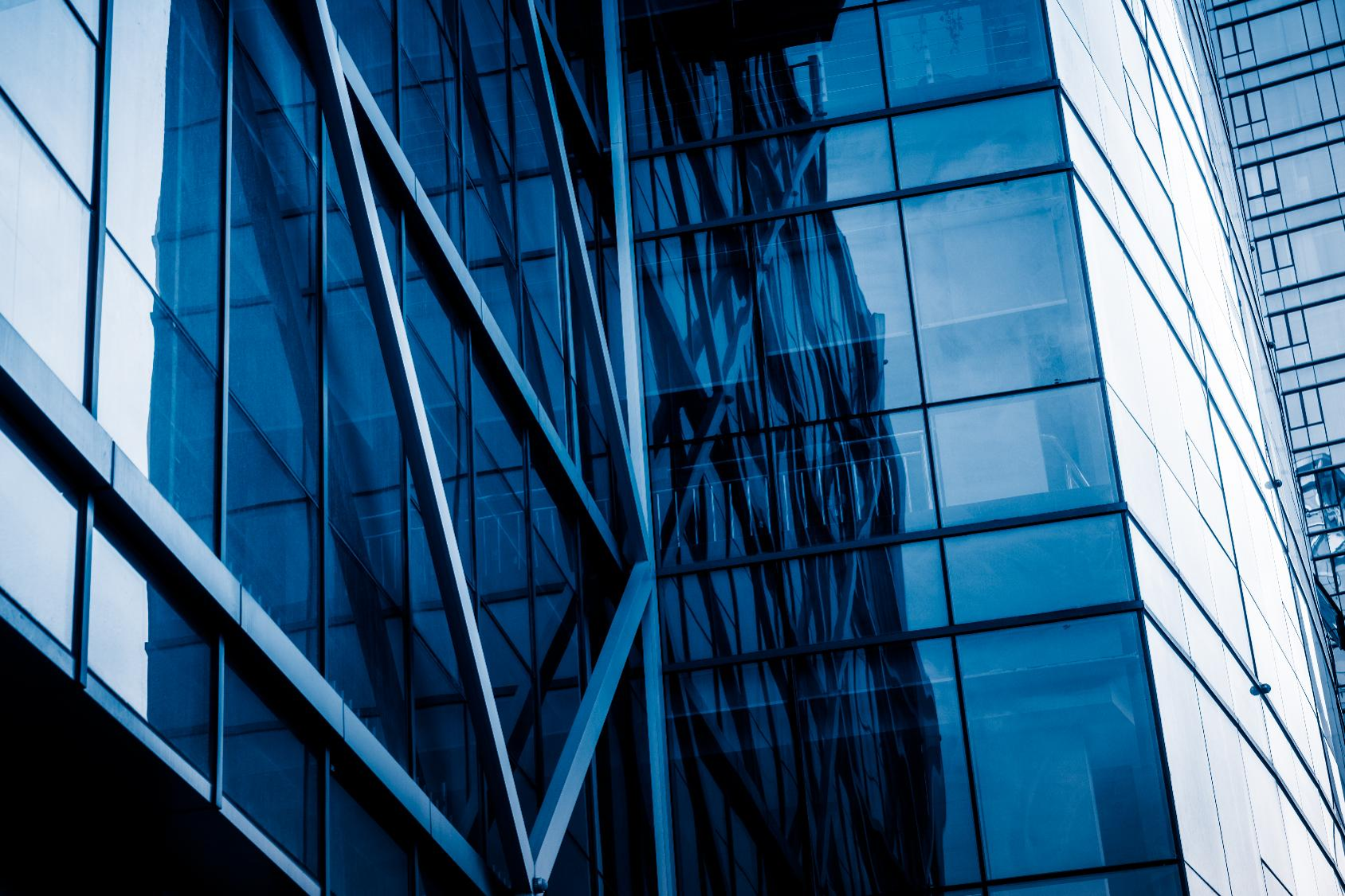 Modern glass office building facade with blue-tinted windows and steel cross-bracing, reflecting surrounding structures.