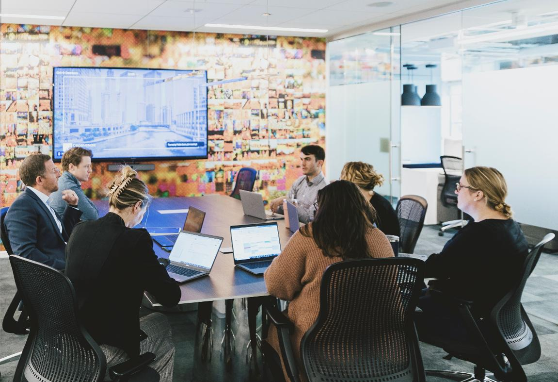 Group of coworkers seated around a conference table in a modern office, using laptops and facing a large screen during a meeting.