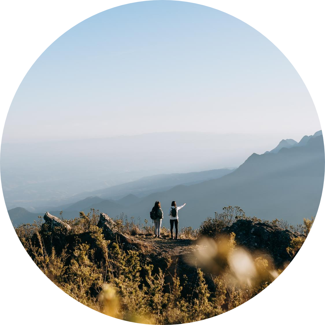 Two hikers standing on a rocky mountain overlook, pointing toward distant layered mountain ranges under a clear sky, viewed through an oval crop.