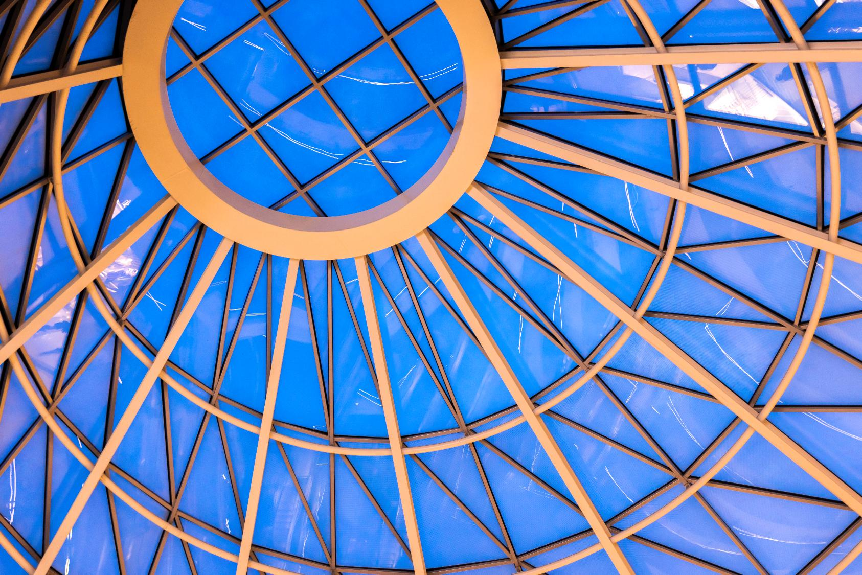 Interior view of a modern glass dome ceiling with radial metal framing and blue-tinted panels forming a circular skylight.